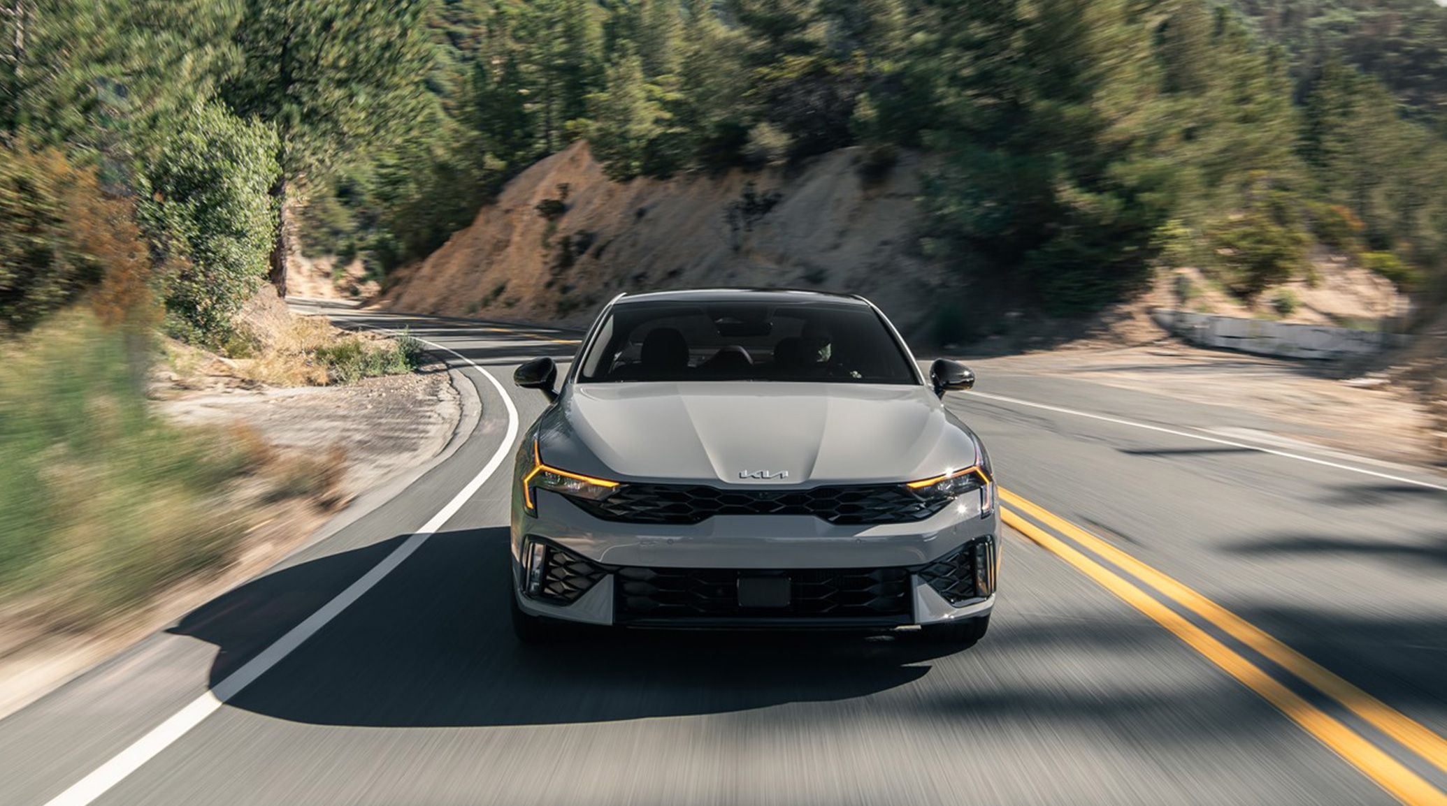 Front view of the 2025 Kia K5 including headlights and grille on a curving mountain road