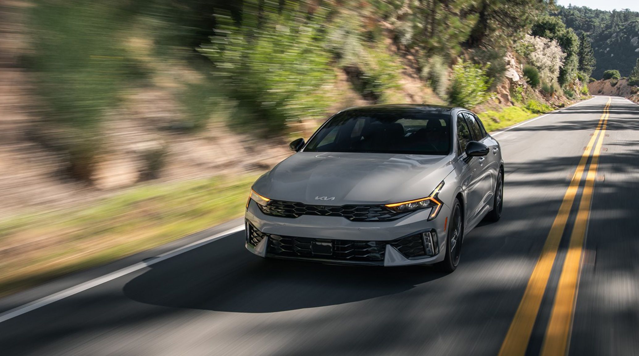 Front driver-side view of the 2025 Kia K5 driving by a blurred mountain background